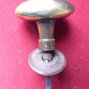 Puede incluir: Un pomo de puerta de latón con una parte superior redonda y abovedada y un largo y delgado vástago de metal. El pomo está unido a una base redonda de metal con un anillo decorativo.