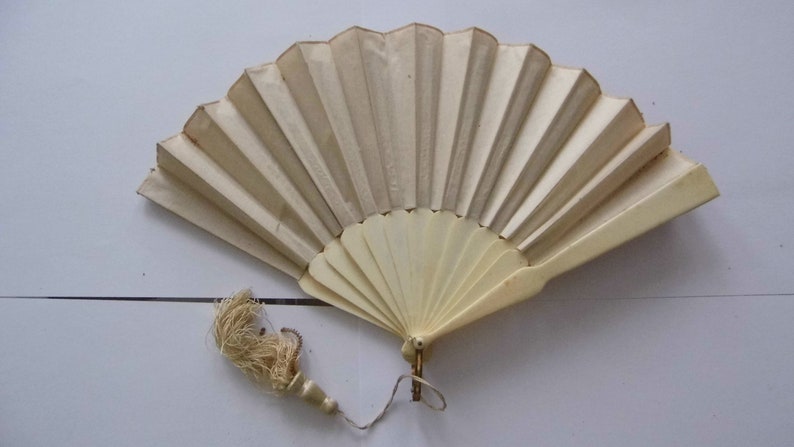 May include: A white folding fan with a cream-colored tassel. The fan is made of paper and has a wooden handle.
