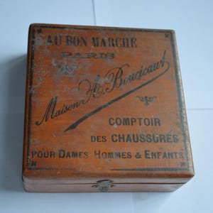 Peut inclure: Une boîte en bois vintage avec un couvercle à charnière et un fermoir en métal. La boîte est teintée d'un brun chaud et comporte des inscriptions noires qui se lisent "AU BON MARCHE PARIS" et "Maison H. Boucicault COMPTOIR DES CHAUSSURES POUR DAMES HOMMES & ENFANTS."