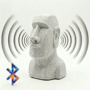 Può includere: Un altoparlante Bluetooth a forma di statua Moai grigia. L'altoparlante ha la forma di una testa con un naso e orecchie grandi. Il simbolo Bluetooth è in basso a sinistra e le onde sonore emanano dall'altoparlante.