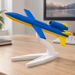 Pode incluir: Um modelo de avião com um esquema de cores azul e amarelo, montado num suporte branco. A aeronave tem um nariz pontiagudo, asas em flecha e uma secção de cauda com estabilizadores verticais e horizontais amarelos sobre uma secretária.