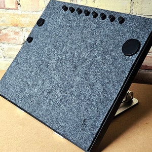 Könnte beinhalten: Ein graues Filz-Schreibtischunterlage mit einem schwarzen Metallständer. Die Unterlage hat einen runden Knopf und mehrere kleine schwarze Knöpfe oben.