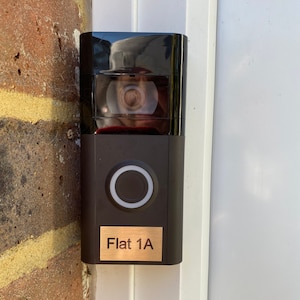 May include: A black doorbell camera with a round button and a gold plaque that reads "Flat 1A".