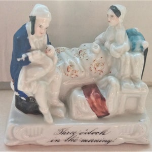 Könnte beinhalten: Eine Porzellanfigur, die eine Szene einer Frau im Bett mit einem Mann zeigt, der ein Baby hält. Die Frau trägt ein weißes Nachthemd und eine schwarze Mütze. Der Mann trägt eine blaue Jacke und eine weiße Hose. Die Figur steht auf einem weißen Sockel mit der Aufschrift "Drei Uhr morgens."