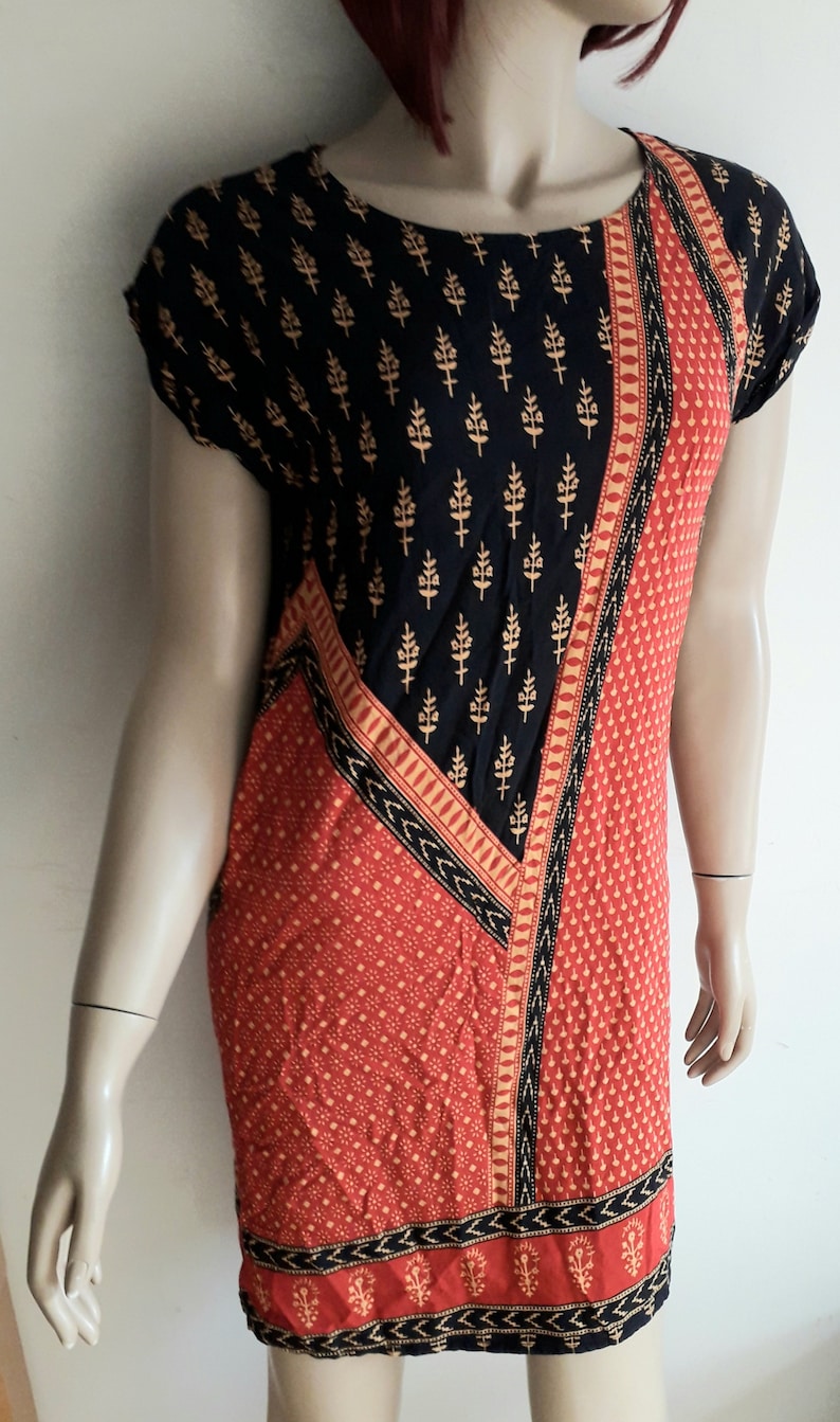 May include: A short-sleeved dress with a black and red geometric pattern. The dress has a black background with a gold floral print in the top half and a red background with a gold dot pattern in the bottom half. The dress has a black and gold trim around the neckline and hem.