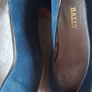 以下が含まれることがあります： ネイビーブルーのスエードパンプス。ポインテッドトゥと茶色の革底。つま先とかかとには、濃い質感の素材を使用。片方の靴の内側にブランド名「BALLY」が見えます。