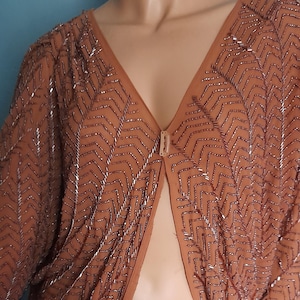 Könnte beinhalten: Braunes, offenes Oberteil mit Perlenbesatz und V-Ausschnitt. Das Oberteil hat ein Zickzackmuster aus silbernen Perlen und einen kleinen Verschluss. Lange Ärmel und transparenter Stoff.