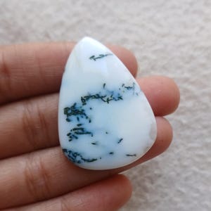 Puede incluir: Una gema blanca en forma de lágrima con inclusiones azul oscuro y negro. La piedra pulida se sostiene en una mano, mostrando su superficie lisa y patrones únicos. La gema es un material natural.