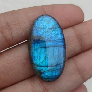 Peut inclure: Une pierre précieuse de labradorite de forme ovale, avec une iridescence bleue vibrante. La pierre polie présente un jeu de couleurs, avec des stries bleues et turquoise. La pierre est tenue dans une main, mettant en valeur sa beauté naturelle et ses motifs uniques.