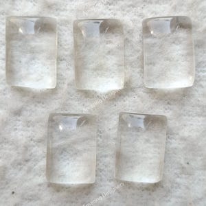 May include: Five clear, rectangular quartz stones are displayed on a light-coloured surface. Each stone has rounded edges and a slightly translucent appearance. The stones are evenly spaced, showcasing their clarity and shape.