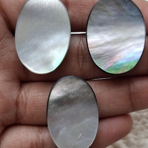 May include: Three oval-shaped mother-of-pearl cabochons. The cabochons have a pearlescent sheen with iridescent colors. The edges of the cabochons are dark. The cabochons are set on pins.