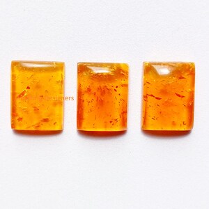 May include: Three rectangular amber-colored cabochons with smooth, polished surfaces and internal golden-orange inclusions. Each piece features a flat base and a slightly domed top, displaying a translucent, warm, honey-toned appearance.