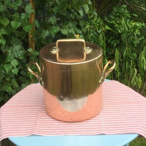 Könnte beinhalten: Ein großer Kupfertopf mit Deckel und zwei Griffen. Der Topf steht auf einem blauen Tisch mit einer rot-weiß gestreiften Tischdecke. Der Topf befindet sich in einem Garten.
