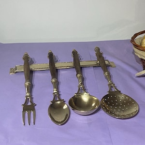May include: Set of four gold-toned kitchen utensils with wooden handles mounted on a wall rack. The utensils include a fork, a spoon, a ladle, and a slotted spoon.