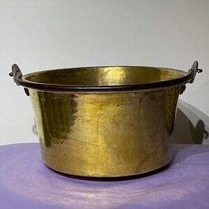 May include: A large, antique brass pot with two iron handles. The pot has a rounded bottom and a wide, shallow opening. The brass has a warm, golden color and shows signs of age and use.