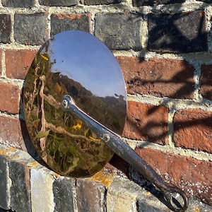 May include: A copper and iron lid with a long handle. The lid is round and has a shiny, reflective surface. The handle is attached to the lid with a metal rivet. The lid is resting on a brick wall.
