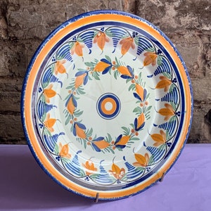 Könnte beinhalten: Ein großer, runder Teller aus Keramik mit weißem Hintergrund und einem blauen und orangefarbenen Blumendesign. Der Teller hat einen erhöhten Rand und einen leicht gewellten Rand.