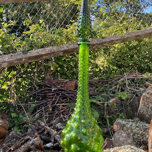 Peut inclure: Une bouteille en verre vert, haute et volumineuse, avec une base bulbeuse et un sommet pointu. La bouteille est recouverte de petites bosses saillantes.