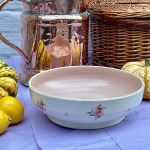 Puede incluir: Un tazón de cerámica blanca con un borde rosa y diseños florales. El tazón está sentado sobre un mantel morado junto a una jarra de cobre y una cesta de mimbre.