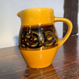 Peut inclure: Un petit pichet en céramique jaune avec un motif de nœud celtique brun et noir. Le pichet a une anse et un corps arrondi.