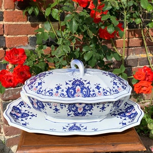 Könnte beinhalten: Eine weiße Keramik-Servierplatte mit einem blauen und rosa floralen Design. Die Platte hat einen Deckel und eine passende Untertasse. Die Platte steht auf einem Holztisch vor einer Backsteinmauer mit roten Rosen.