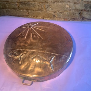 May include: A round copper pan with a handle. The pan has a sun design with rays emanating from the center and two fish designs below the sun.