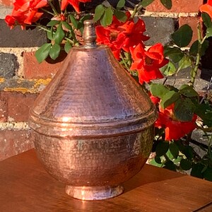 May include: A hammered copper vase with a rounded, bulbous shape and a domed lid. The vase is sitting on a wooden surface in front of a brick wall with red roses.