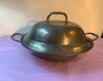 May include: A vintage, pewter-toned metal serving dish with a domed lid and two side handles. The lid has a decorative handle. The dish has a round base and is likely used for serving food. The metal has a slightly aged appearance.