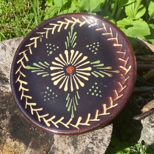 Puede incluir: Un plato de cerámica marrón con un diseño decorativo de una flor estilizada en amarillo, verde y marrón. El plato tiene un esmalte marrón oscuro y un borde blanco.