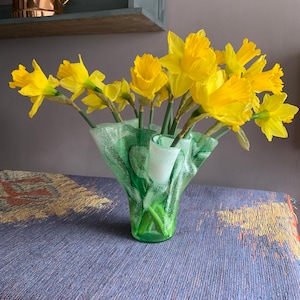 May include: A bouquet of yellow daffodils in a green and white glass vase with a ruffled edge. The vase is sitting on a blue and brown patterned tablecloth.