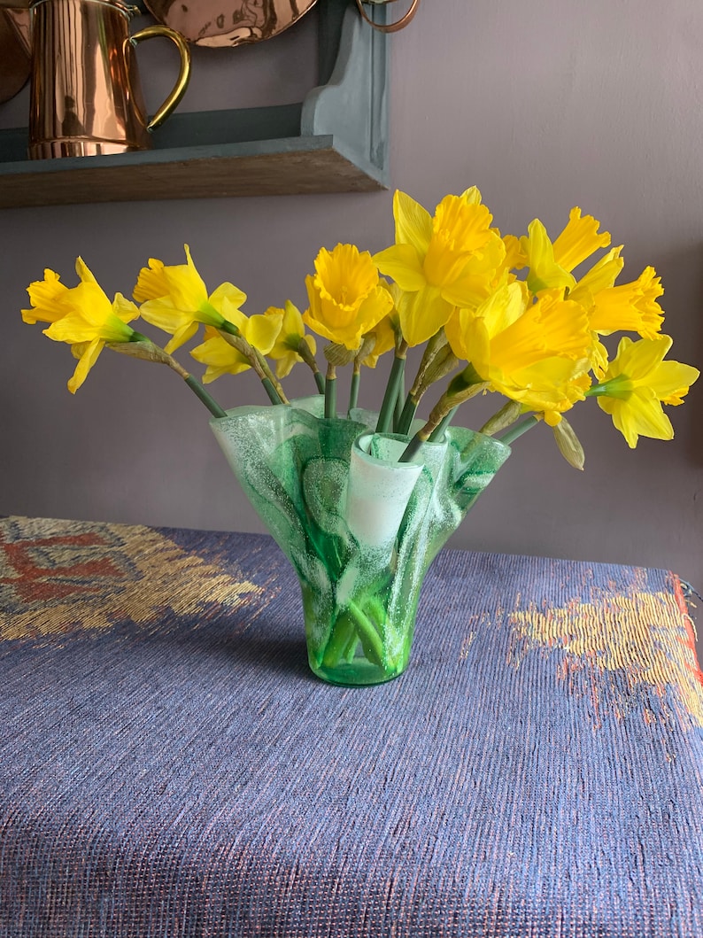May include: A bouquet of yellow daffodils in a green and white glass vase with a ruffled edge. The vase is sitting on a blue and brown patterned tablecloth.