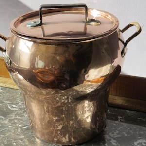 Op de afbeelding: Een grote koperen pot met een deksel en twee handvatten. De pot is glanzend en heeft een gehamerde afwerking. Hij staat op een houten dienblad met een marmeren oppervlak.