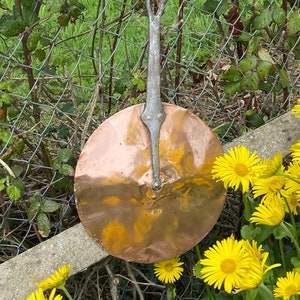 May include: A copper pan with a long, silver handle. The pan is hanging on a fence with yellow flowers in the foreground.