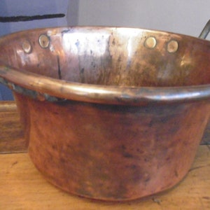 Puede incluir: Una olla de cobre vintage con dos asas de latón. La olla tiene un fondo redondeado y una abertura ancha y poco profunda. El cobre tiene una pátina de edad y desgaste.