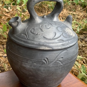May include: A black ceramic pitcher with a handle and a spout. The pitcher has a floral design and the word "Velez" inscribed on the side.