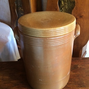 以下が含まれることがあります： 蓋付きの茶色の陶器の壺。壺は表面がざらざらしていて、木のテーブルの上に置かれています。