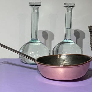 Pode incluir: Uma panela de cobre com uma alça longa está sobre uma superfície roxa ao lado de duas garrafas de vidro transparente.