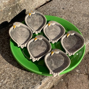 Op de afbeelding: Zes keramische schaaltjes in de vorm van vogels, elk met een grijze body, witte details en een gele snavel. Ze zijn gerangschikt op een felgroene schaal, wat een artistieke presentatie creëert. Perfect voor het serveren van snacks.