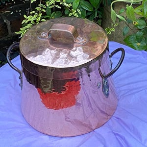Op de afbeelding: Een grote koperen pot met een deksel en twee handvatten. De pot heeft een gehamerde afwerking en staat op een paarse doek.