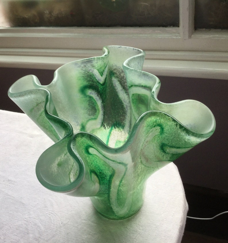 May include: A green and white glass vase with a ruffled, wavy design. The vase is made of clear glass with green and white swirls.