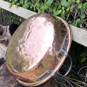 May include: A large, round copper pot with two handles. The pot has a hammered finish and is resting on a wooden surface.