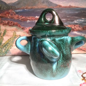 May include: A green and blue ceramic pot with a lid and two handles. The pot has a textured surface and a glossy finish.