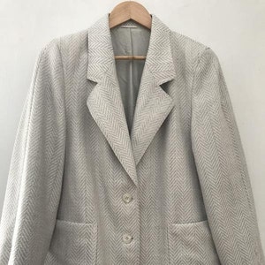Puede incluir: Un blazer beige claro con un patrón de espiga. El blazer tiene solapas con muescas, dos bolsillos delanteros y dos botones a juego. La chaqueta está colgada en una percha de madera.