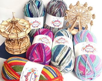 May include: Six balls of yarn in various colors and patterns, including blue, green, pink, purple, and yellow. The yarn is labeled "Fairground" and is arranged on a white surface with a wooden carousel and Ferris wheel in the background.