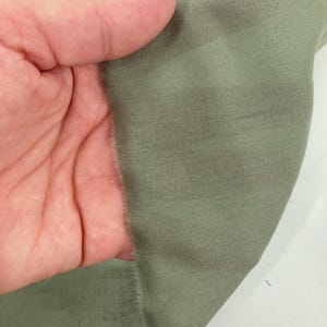 May include: A piece of sage green fabric with a visible woven texture and a raw, frayed edge. The material displays a consistent muted green tone throughout, with a subtle sheen and a fine, tight weave pattern visible across the surface.