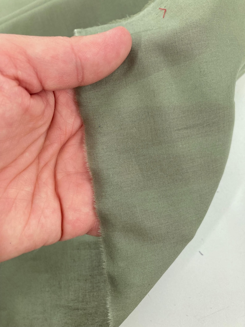 May include: A piece of sage green fabric with a visible woven texture and a raw, frayed edge. The material displays a consistent muted green tone throughout, with a subtle sheen and a fine, tight weave pattern visible across the surface.