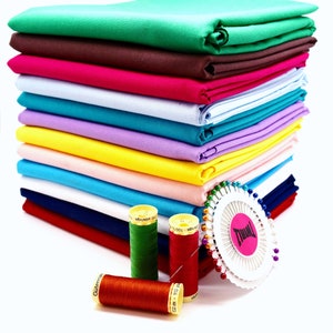 May include: A stack of colourful fabric swatches in various shades of green, brown, pink, blue, purple, yellow, and peach. The fabric is folded neatly and arranged in a pile. There are also spools of thread and a pincushion with pins.