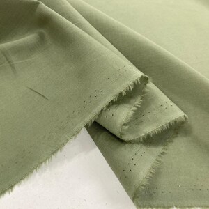 May include: A length of sage green fabric with a soft, matte texture. The material is draped to show folds and creases, revealing a raw, frayed edge along the selvage. The selvage features a distinct row of small, evenly spaced pinhole perforations.