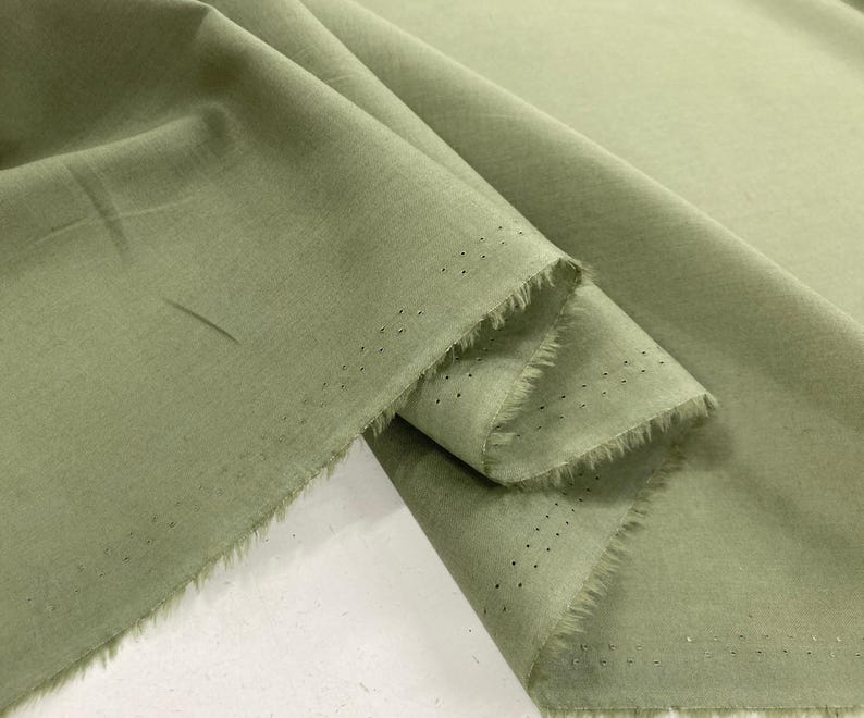 May include: A length of sage green fabric with a soft, matte texture. The material is draped to show folds and creases, revealing a raw, frayed edge along the selvage. The selvage features a distinct row of small, evenly spaced pinhole perforations.