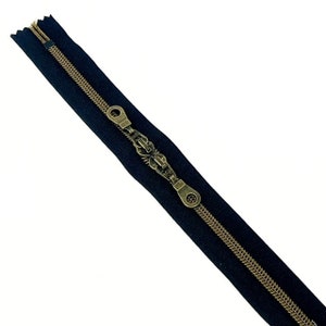 May include: A black zipper with a brass slider and pull. The zipper is closed and the slider is in the center of the zipper.